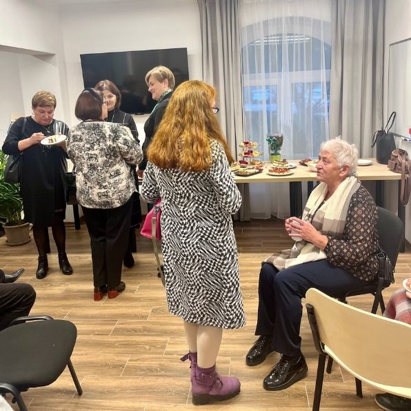 dwie panie rozmawiające (jedna siedząca, druga stojąca), w tle inni ludzie